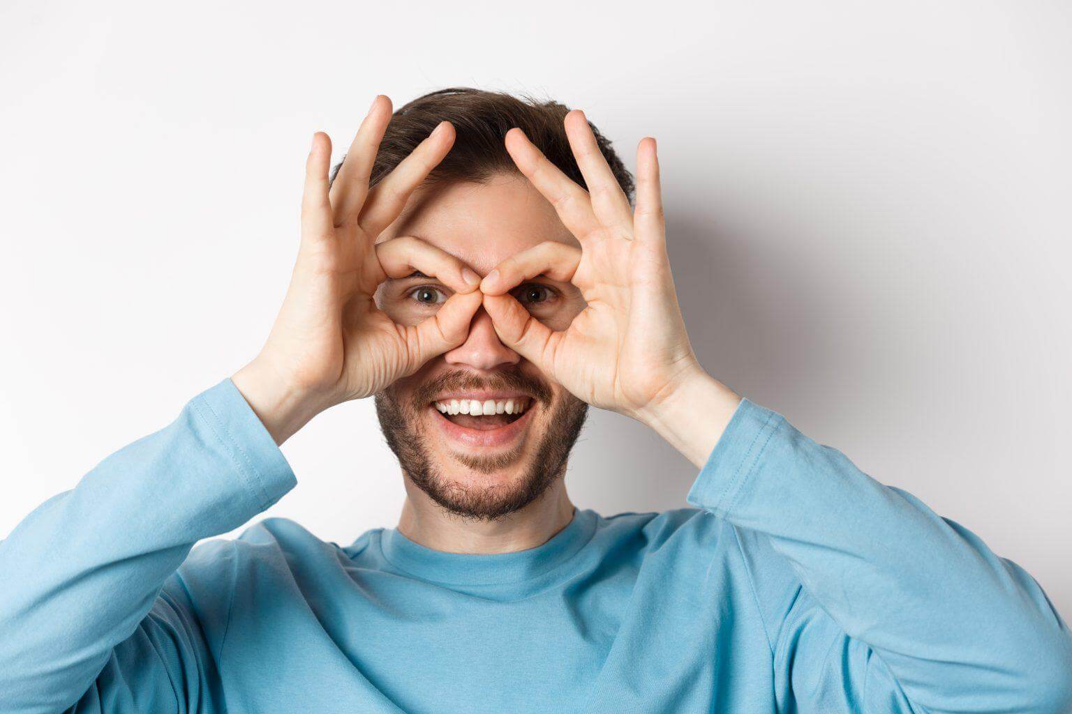 Happy smiling man looking through hand binoculars with amazed face, checking out promo offer, standing on white background